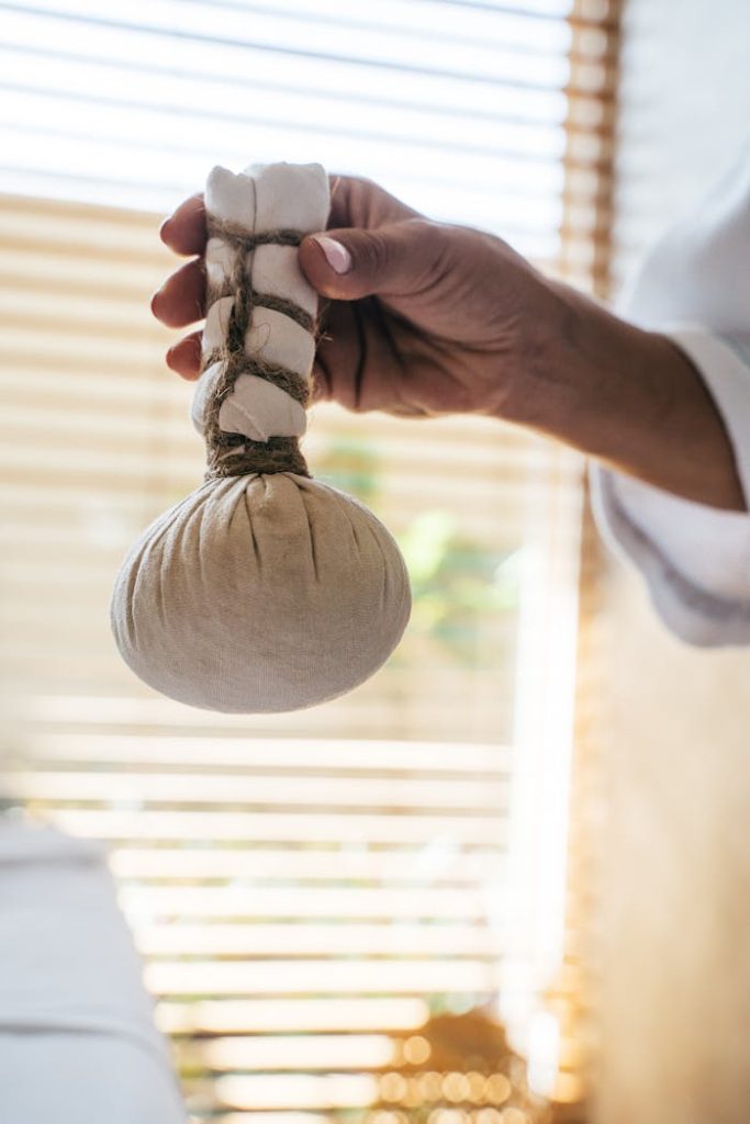 A hand holding a herbal massage compress in a serene indoor setting with soft lighting.